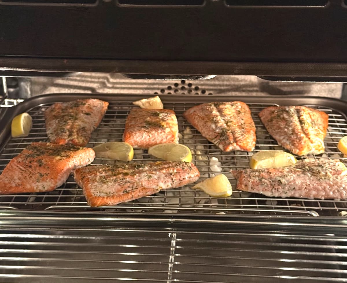 Six salmon fillets seasoned with herbs and lemon wedges, broiling on a wire rack in the oven