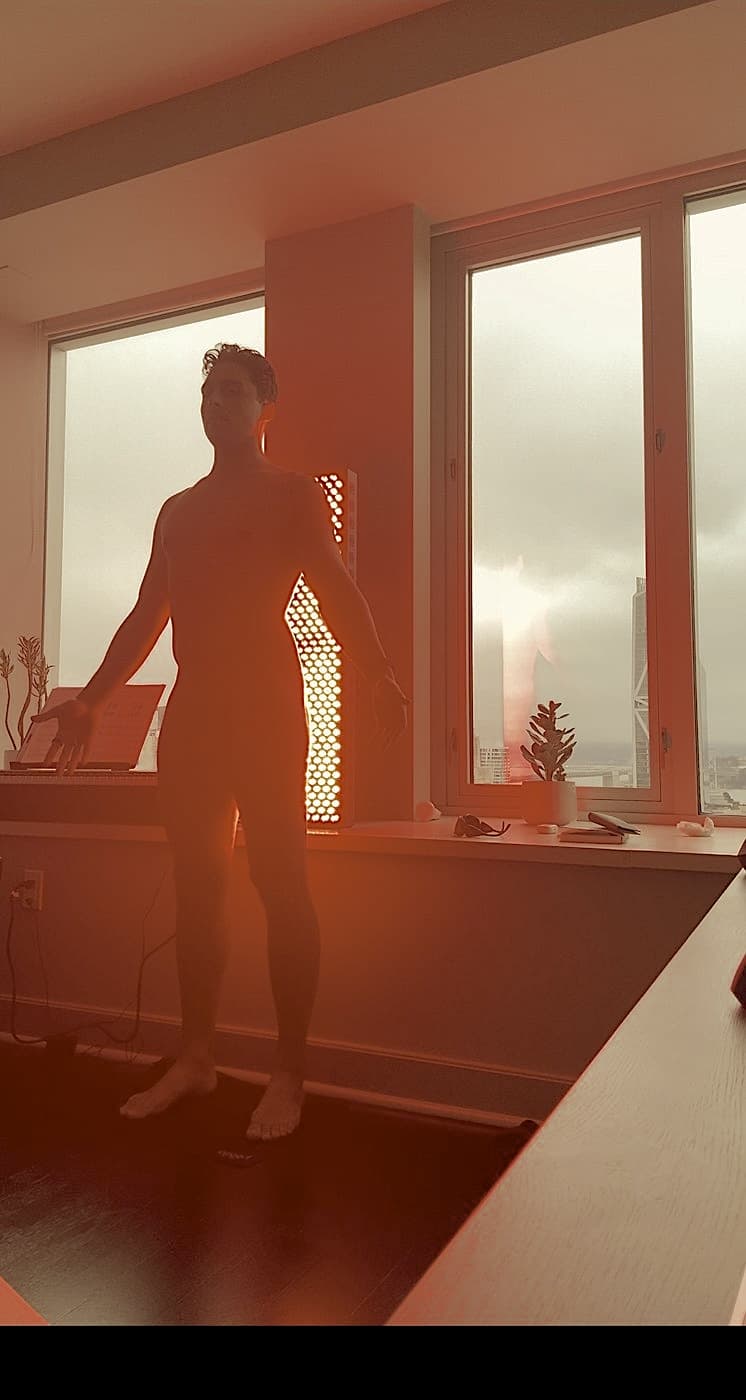 Nick standing in front of a tall red light therapy panel by a window overlooking the city, the room bathed in red